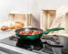 A green frying pan with tomatoes sits on a black induction stovetop, with bread and wooden crates in the background. Produktbild-Vorschau