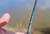 A close-up shot of a fishing rod with a fishing line, held by a person's thumb, with a body of water in the background. Produktbild-Vorschau