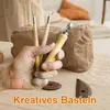 A hand holding pottery tools in front of a large block of clay. Produktbild-Vorschau