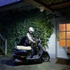 A person in a helmet is about to get on a scooter parked in front of a house with ivy-covered walls and a door. Produktbild-Vorschau
