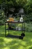 A black serving cart is set up in a garden with drinks and glasses on it. Produktbild-Vorschau