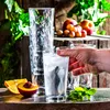 A hand is reaching for a glass of iced water, with a stack of glasses, fruit, and greenery in the background. Náhled obrázku produktu