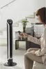 A woman is using a remote control to operate a tall black and silver tower fan in a living room. Produktbild-Vorschau