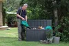 A man is using a leaf blower to clean out a garden storage box in a backyard. Anteprima dell’immagine prodotto