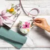 A hand is placing a small sprig of flowers into a mug with a floral design, next to a gift box and a folded napkin. Podgląd zdjęcia produktu