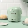 A sage green mini rice cooker is shown on a light green surface, with cooked rice in small bowls on either side. The background is white subway tile. Produktbild-Vorschau
