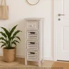 A narrow, white-washed wooden cabinet with four drawers stands in a hallway next to a white door. A green plant in a woven basket is to the left of the cabinet. Podgląd zdjęcia produktu