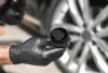 A gloved hand holds a black applicator brush with black bristles, with a car wheel in the blurred background. Anteprima dell’immagine prodotto