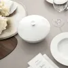 A table setting with a white lidded bowl, flowers, a glass of water, and a menu. Produktbild-Vorschau