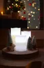A festive Christmas arrangement with LED candles and miniature trees on a wooden slice, with a blurred Christmas tree and lights in the background. Produktbild-Vorschau