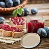A rustic scene featuring plum jam on toast, a jar of jam, and fresh plums, with a lid in the foreground. Produktbild-Vorschau