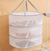 A three-tiered mesh drying rack hanging against a light orange wall, with a white clothing rack visible on the right. Produktbild-Vorschau