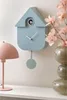 A light blue cuckoo clock is displayed on a wall, next to a pink lamp and a vase with pink flowers. The clock has a pendulum and a small bird visible. Náhled obrázku produktu