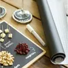 A close-up shot of a small blackboard with food items and a wine glass on a wooden surface. A roll of black paper and a grid paper are also visible. Produktbild-Vorschau