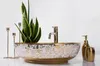 A decorative washbasin with a gold faucet and ornate gold and white patterns, placed on a white surface with decorative gold candle holders and soap dispensers. Produktbild-Vorschau