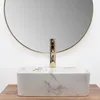 A close-up of a marble-patterned vessel sink with a gold faucet, set against a circular mirror and a wooden surface. Podgląd zdjęcia produktu