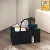 A dark gray felt shopping bag filled with papers and boxes sits on a wooden floor next to a white cabinet with a framed picture of a baby. Produktbild-Vorschau