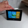 A hand is touching a digital weather station displaying outdoor and indoor temperature, humidity, and weather forecast. Produktbild-Vorschau