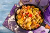 A close-up overhead shot of a pan filled with a colorful vegetable stir-fry, placed on a blue wooden surface with decorative purple and gold marbled paper underneath. Náhled obrázku produktu
