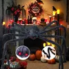 A large, fuzzy black spider decoration with red eyes is displayed in front of a fireplace decorated for Halloween, with pumpkins, skulls, and other spooky items. Produktbild-Vorschau