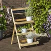 A wooden plant stand with three shelves, holding potted plants, on a wooden deck with a brick wall and greenery in the background. Produktbild-Vorschau