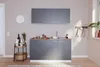 A modern gray kitchenette with a wooden countertop, sink, stove, and cabinets, set against a light pink wall with a clock and a small serving cart with bottles and a basket. Produktbild-Vorschau