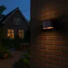 A modern outdoor wall light illuminates a brick wall next to a house and garden at dusk. Produktbild-Vorschau