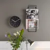 A wall with a clock and a magazine rack, with flowers and a lamp in the foreground. Produktbild-Vorschau