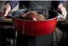 A person is holding a red cast iron pot with a roasted chicken inside, steam rising from the pot. Produktbild-Vorschau