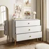 A white dresser with three drawers is placed in a room with artwork on the wall and a large mirror. Produktbild-Vorschau