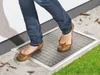 A person is stepping onto a metal grate designed to cover a basement window well, with grass and leaves visible. Produktbild-Vorschau