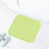A light green square bath mat with suction cups lies on a marble-tiled floor next to a white bathtub. Two light blue flip-flops are placed to the left of the mat. Produktbild-Vorschau
