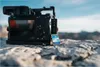 A camera and an SD card are placed on rocks outdoors with a blurred background of mountains and sky. Podgląd zdjęcia produktu