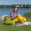 A woman is sitting by a lake, using her phone which is connected to a power bank. Produktbild-Vorschau