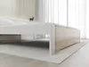A low-angle shot of a bed frame with white sides and a light wood-grain base, set against a backdrop of vertical wooden slats and sheer curtains. Náhled obrázku produktu