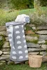A gray blanket with a sheep pattern and a sheep-shaped pillow are placed on a stone wall outdoors, with a basket nearby. Produktbild-Vorschau