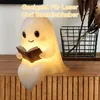 A cute ghost-shaped night light is sitting on a wooden surface, reading a book. There is a decorative basket with dried plants in the background. Produktbild-Vorschau