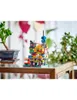 A small, colorful LEGO model of a building is displayed on a wooden table, with plants in the background. Náhled obrázku produktu