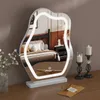 A vanity mirror with LED lighting is placed on a wooden desk, reflecting a bedroom scene. Two candle holders with lit candles are to the right of the mirror. Produktbild-Vorschau