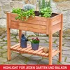 A wooden raised garden bed filled with plants, placed on a stone patio. A watering can and a potted flower are on the shelf below. Produktbild-Vorschau