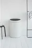 A white Brabantia Touch Bin Mülleimer with a black lid stands on a white wooden floor against a white wall. A black stool with a white pitcher is to the left, and a black towel rack with a black and white polka dot towel is to the right. Voir les images du produit