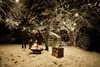 People gathered around a fire pit in a snowy backyard at night, with string lights and a greenhouse in the background. Podgląd zdjęcia produktu