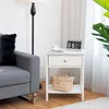 A white nightstand with a drawer and a shelf sits next to a gray sofa. A black floor lamp is behind the sofa. A woven basket is on the shelf of the nightstand, and a cup and saucer and a flip clock are on top of the nightstand. A large green plant is visib Anteprima dell’immagine prodotto