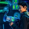 A young boy is holding a toy blaster that is emitting smoke, in a dimly lit room with colorful lights in the background. Náhled obrázku produktu