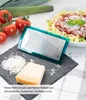 A close-up of a grater insert and cheese on a dark slate board, with tomatoes and leeks in the background, and a bowl of pasta with sauce and grated cheese in the upper right corner. Produktbild-Vorschau