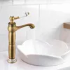 A close-up shot of a retro-style bronze faucet with a white handle, dispensing water into a decorative white sink. The background features white subway tiles. Podgląd zdjęcia produktu