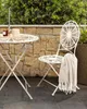 A white metal bistro set on a stone patio with greenery in the background. Produktbild-Vorschau