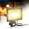 A floodlight with a motion sensor is shown in a dark outdoor setting, with a brick wall and foliage in the background. Náhled obrázku produktu