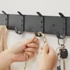 A close-up shot of a black wall-mounted coat rack with several hooks. Hands are shown attaching a gold chain purse to one of the hooks, and a set of keys is hanging from another hook. Produktbild-Vorschau
