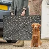 A person is holding a folded dog bed next to a car, with a dog sitting in front of the bed. Produktbild-Vorschau